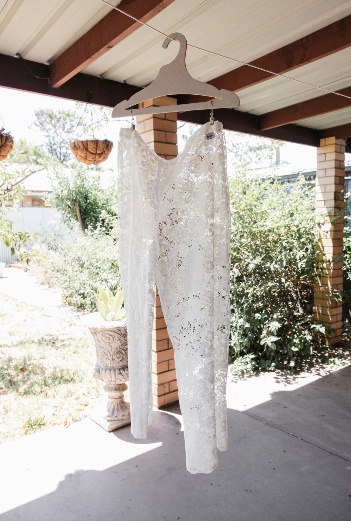 White lace wedding dress hanging on a wooden hanger outdoors.