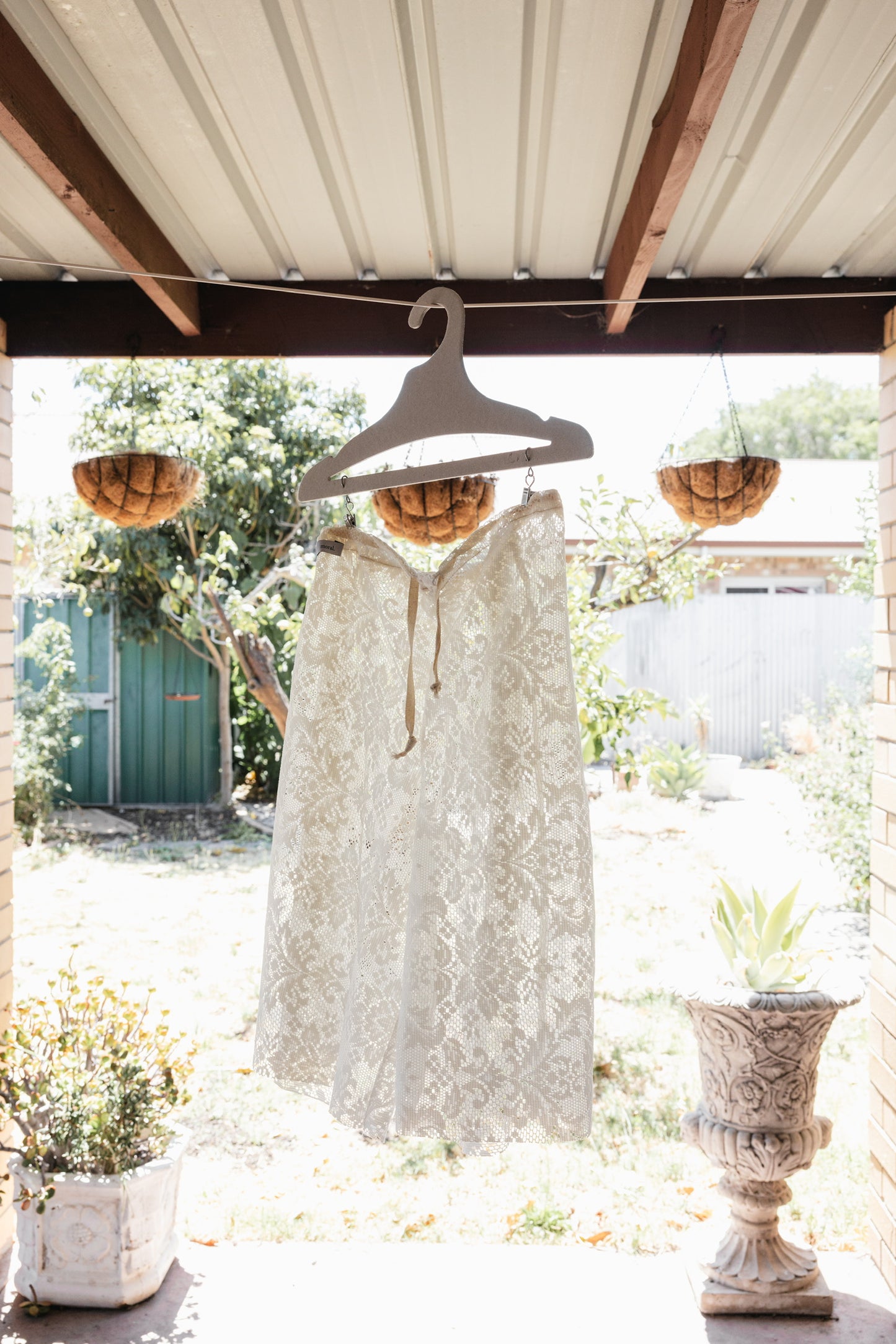 White lace dress hanging on a wooden hanger under a covered outdoor area with plants and flowers.