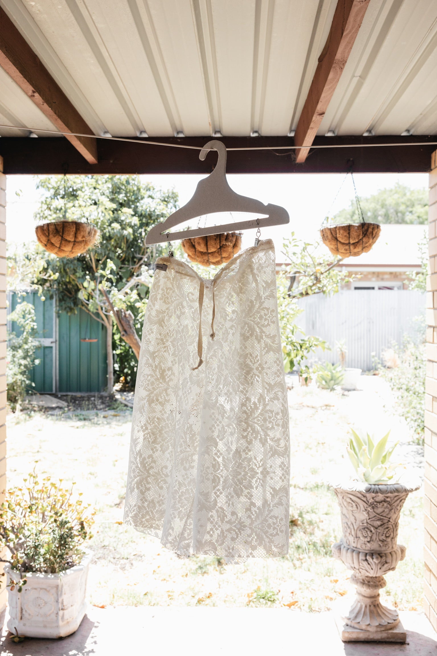 White lace dress hanging on a wooden hanger under a covered outdoor area with plants and flowers.