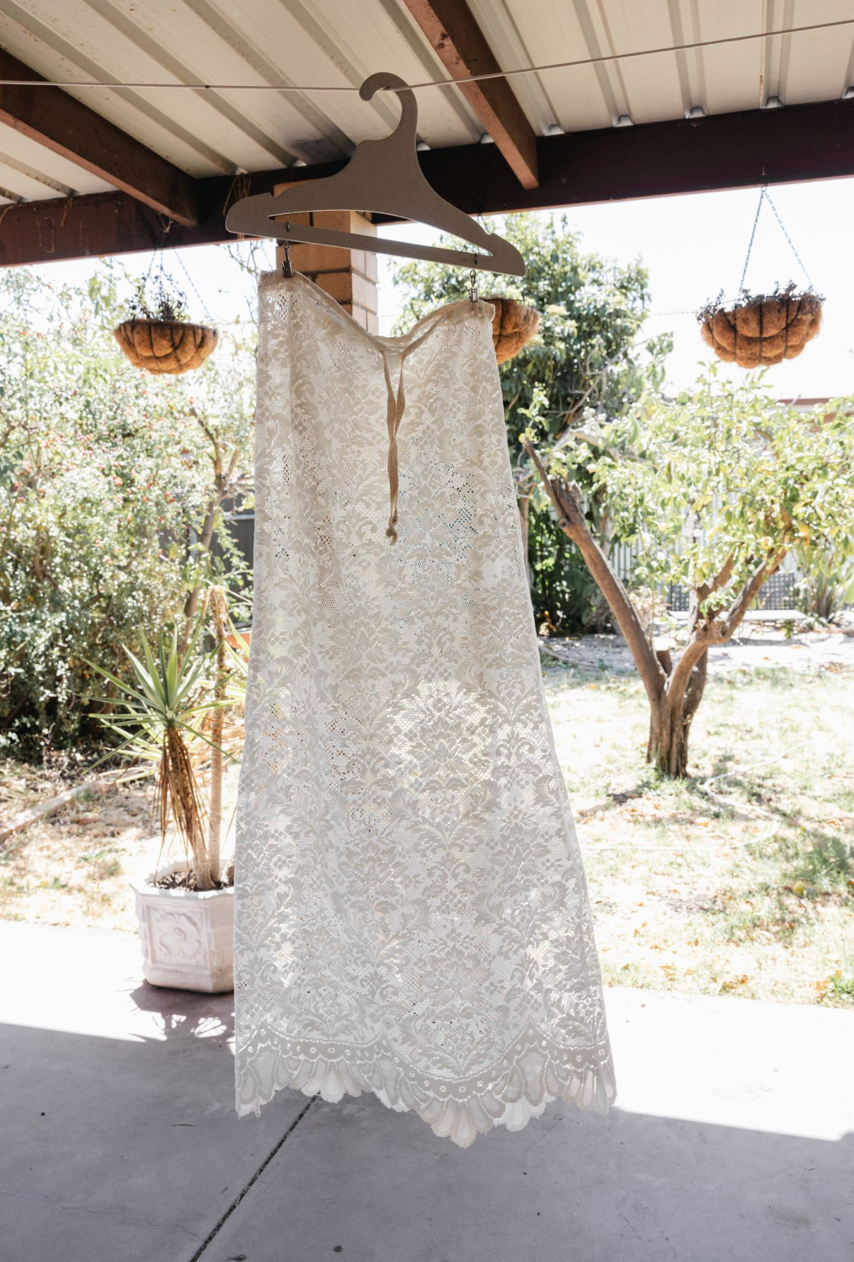 White lace wedding dress hanging on a wooden hanger outdoors.
