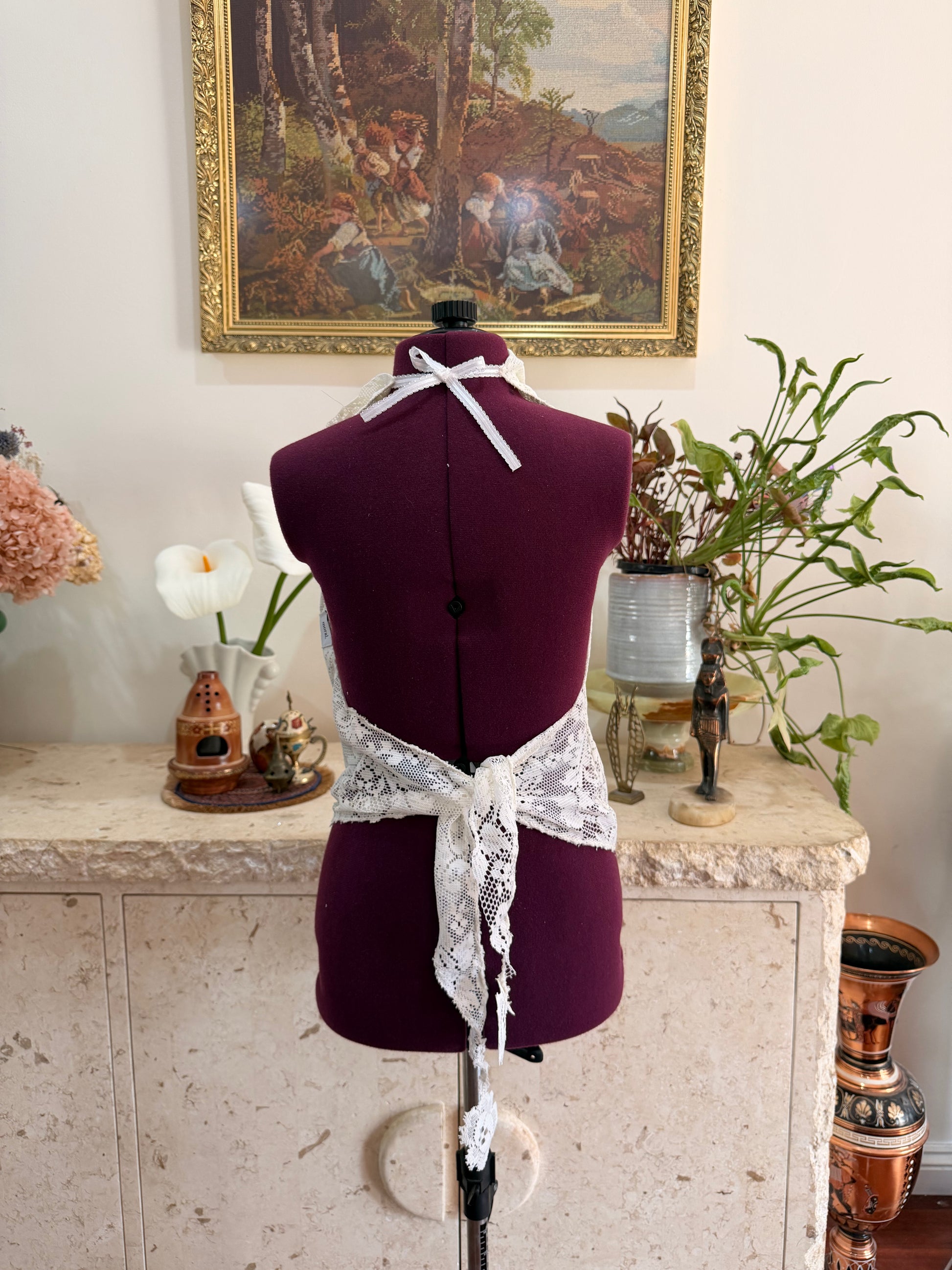A dark plum purple fabric dress form mannequin with a white lace apron tied around its waist, set against a vintage interior background with a painting and plants.