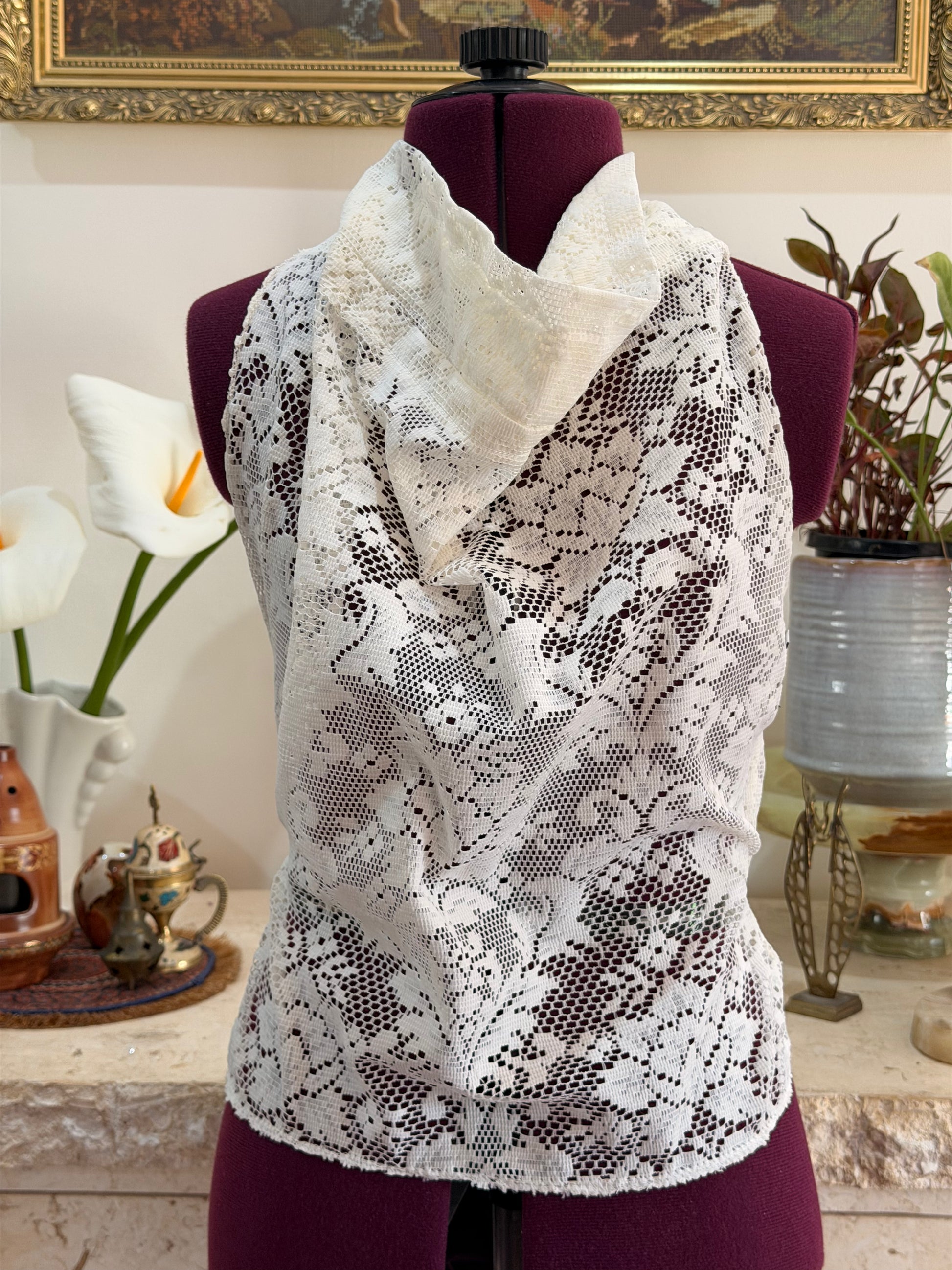 White floral lace cowl neck top displayed on a burgundy dress form mannequin, with a matching white lace shawl draped over the shoulders, set against a home interior background with a framed picture and potted plants.